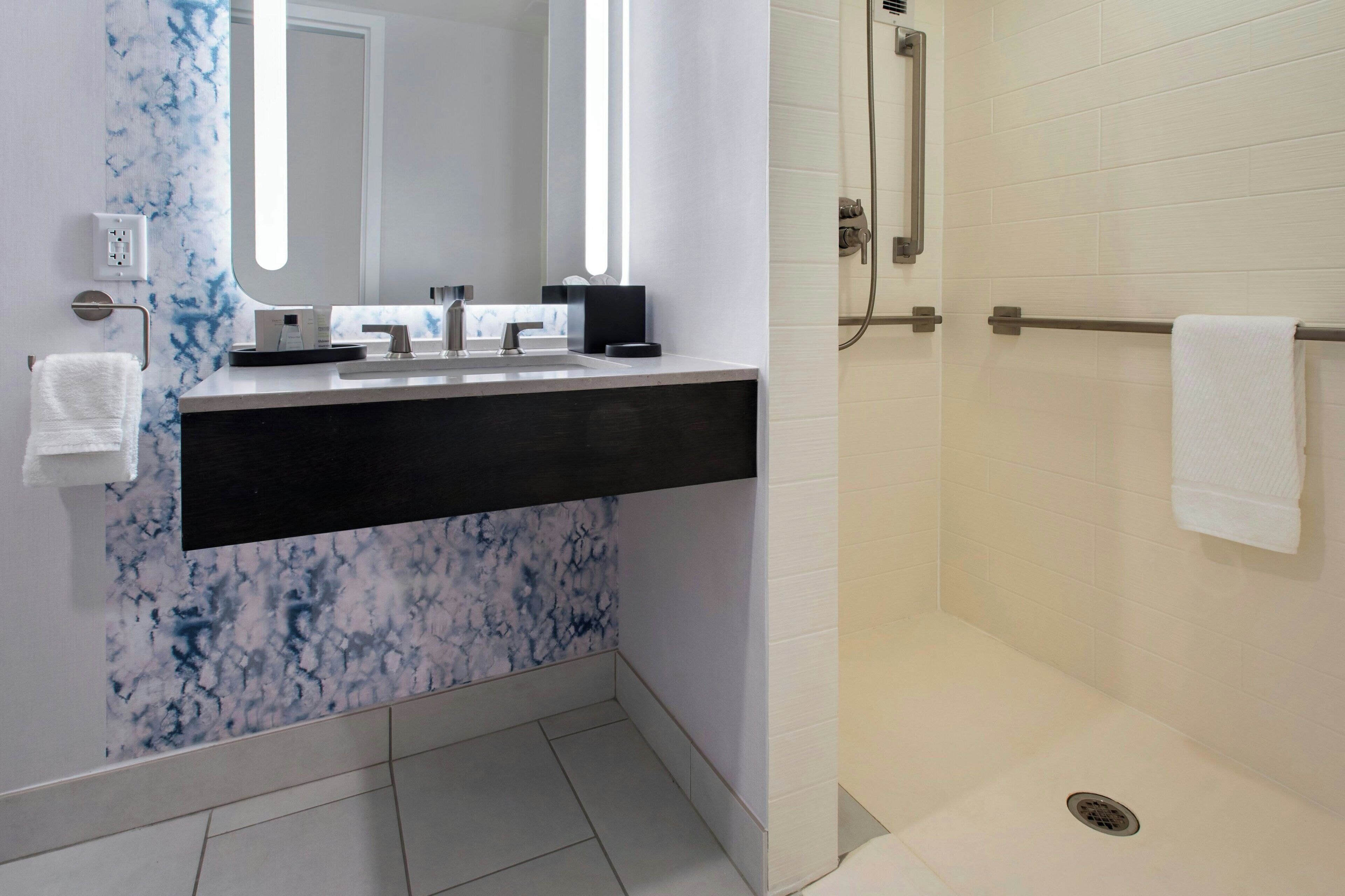 A modern bathroom with a marble backsplash, a black countertop, a large mirror, and a shower area.