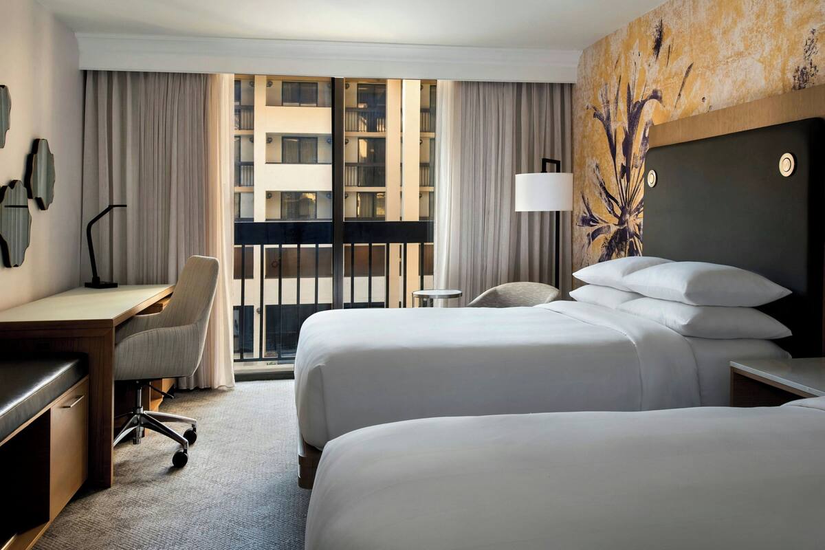 A hotel room with two beds, a desk, a chair, a lamp, and a view of a building through the window.