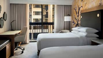A hotel room with two beds, a desk, a chair, a lamp, and a view of a building through the window.