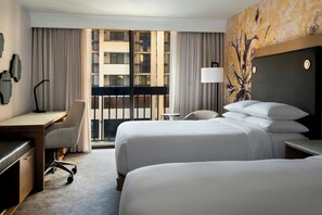 A hotel room with two beds, a desk, a chair, a lamp, and a view of a building through the window.