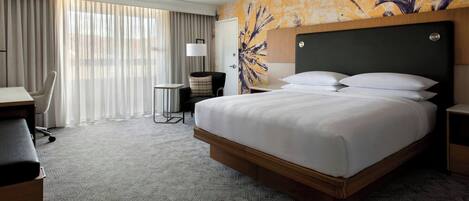 A hotel room with a large bed, a desk, a chair, and a view of the outdoors through the window.