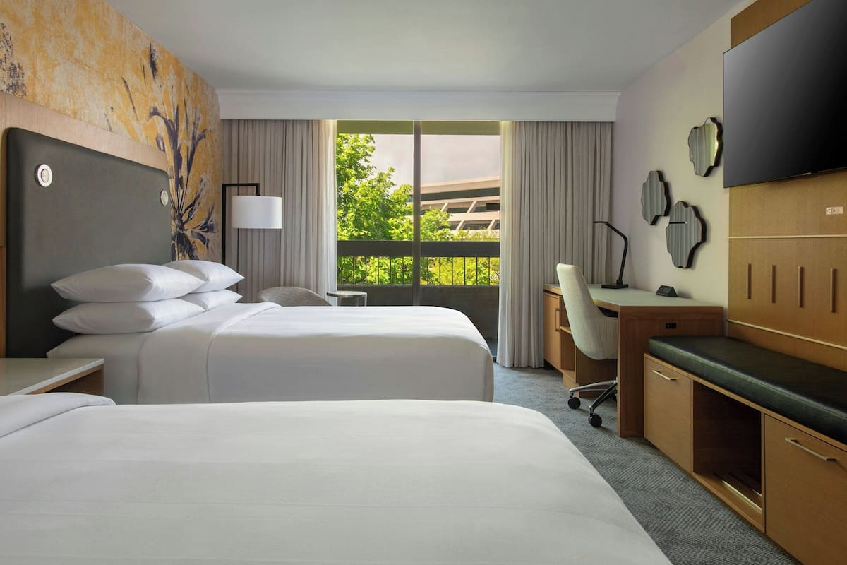 A hotel room with two beds, a desk, a chair, a TV, and a view of greenery outside.