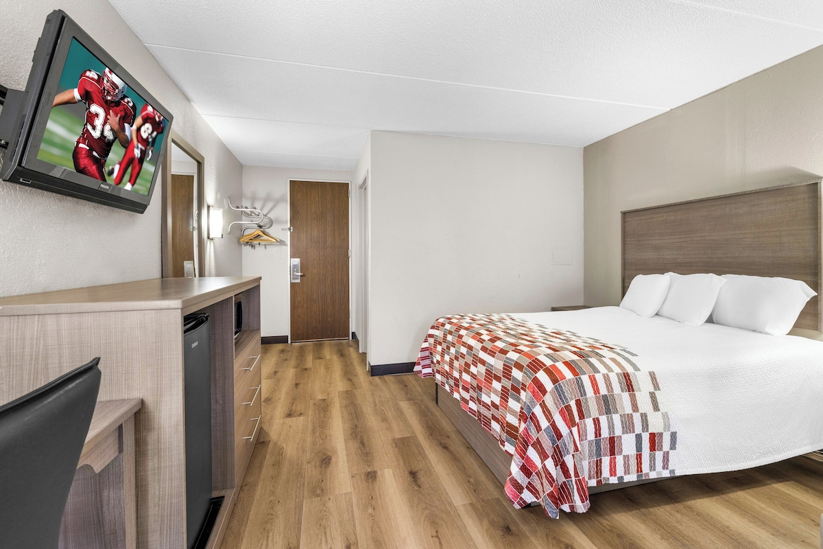 A hotel room with a bed, a TV mounted on the wall, a desk with a chair, and a wooden floor.