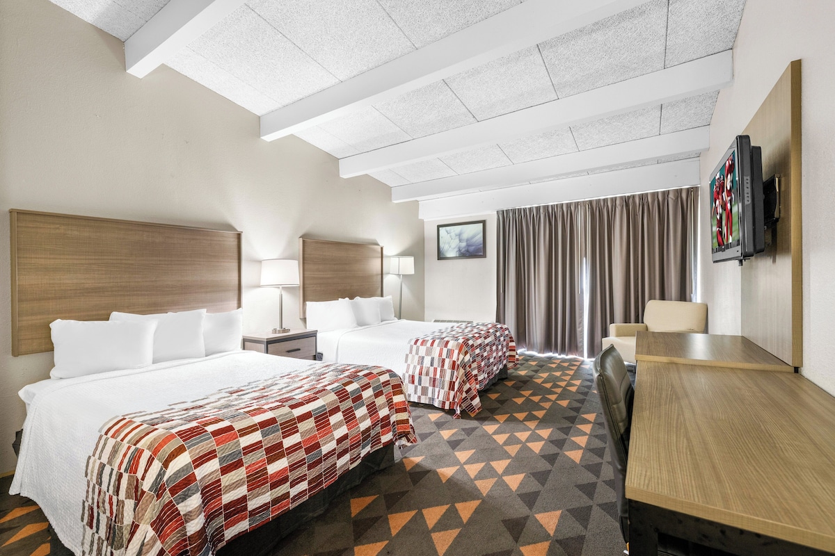 A hotel room with two beds, a desk, a TV, and a window with curtains.