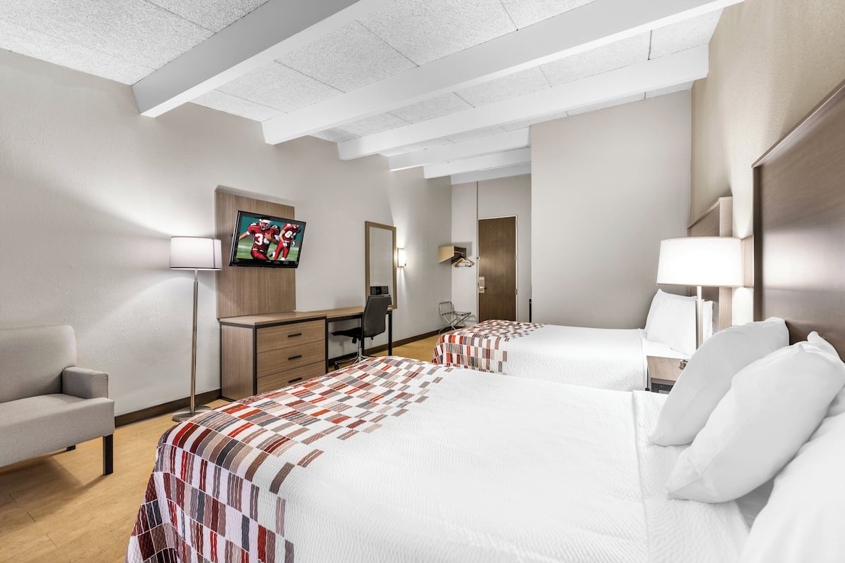 A hotel room with two beds, a desk, a chair, a TV, and a lamp.