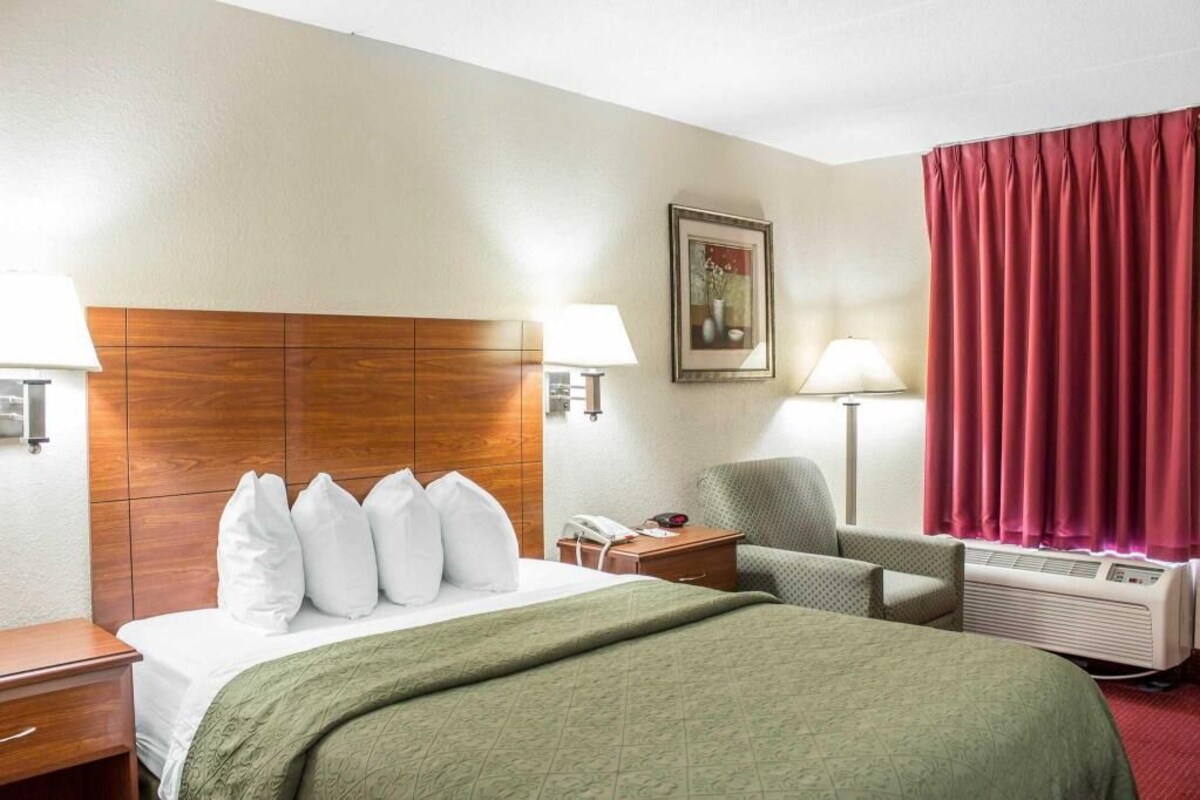 Una habitación de hotel con una cama, dos mesitas de noche, una silla, un escritorio, una lámpara, un cuadro en la pared y una ventana con cortinas.