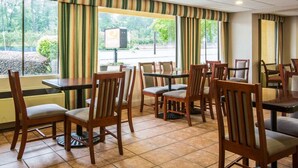 Un restaurante con sillas y mesas de madera, ventanas amplias con cortinas y vistas al exterior con vegetación.