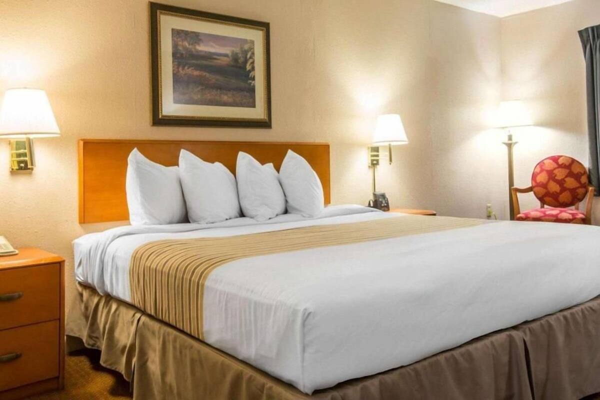 A hotel room with a bed, two bedside lamps, a nightstand, a chair, and a framed picture on the wall.