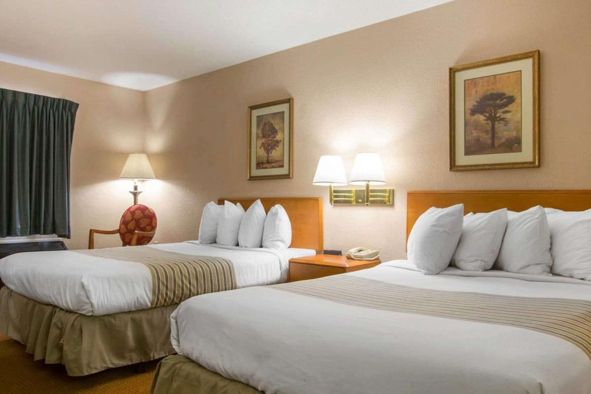 A hotel room with two beds, a desk, a chair, lamps, and two framed pictures on the walls.