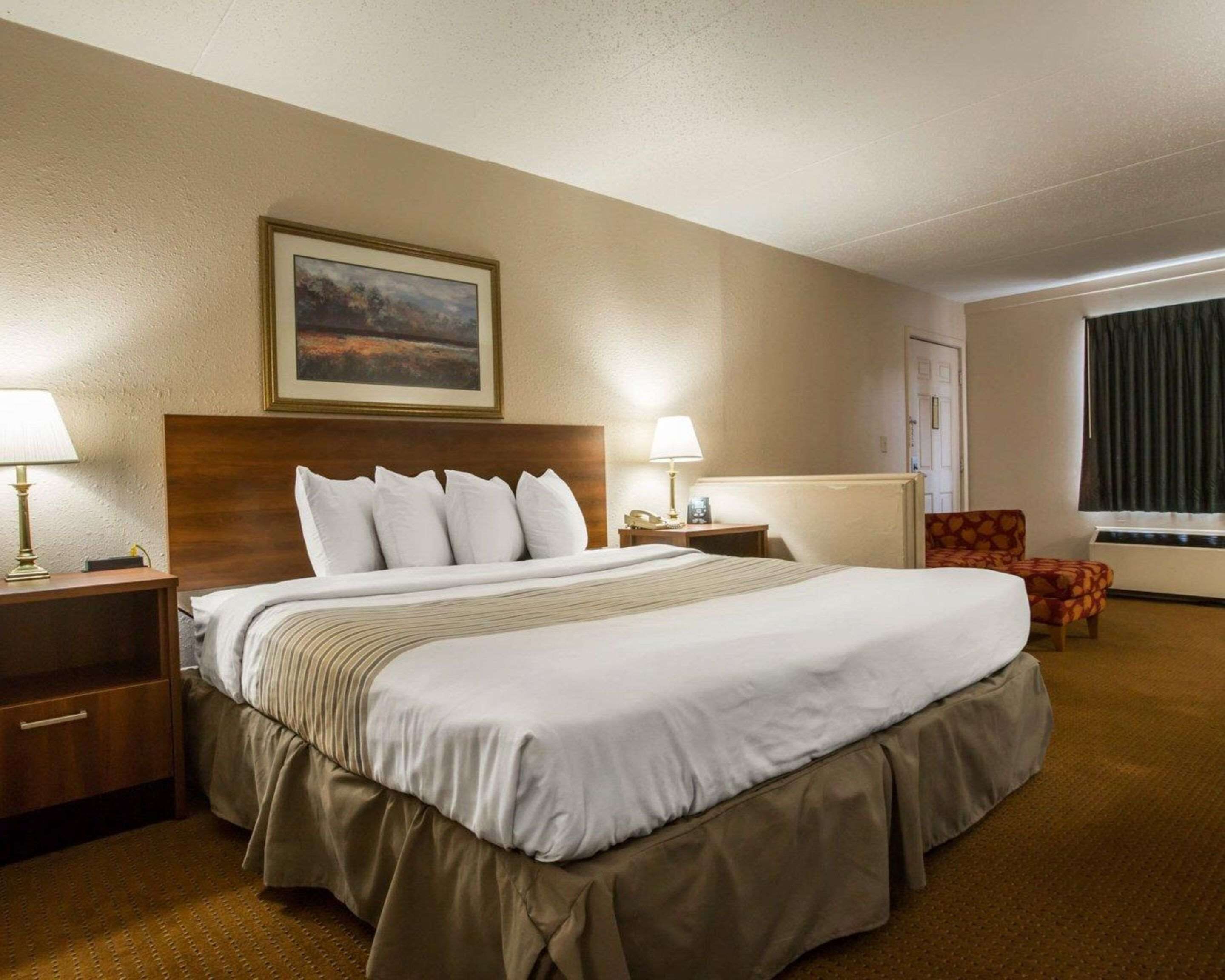 Ein Hotelzimmer mit einem großen Bett, zwei Nachttischen inklusive Lampen, einem Sessel, einem Bild an der Wand und einem Fenster mit Vorhängen.