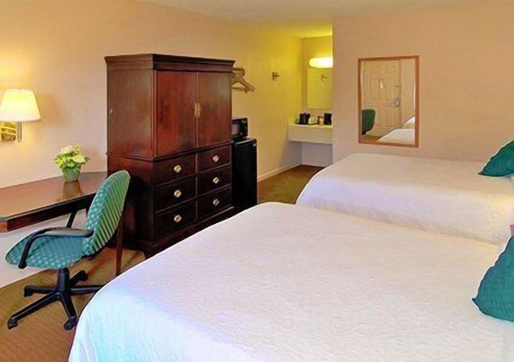 Habitación de hotel con dos camas, un escritorio con silla, un armario, una lámpara y un baño con espejo.