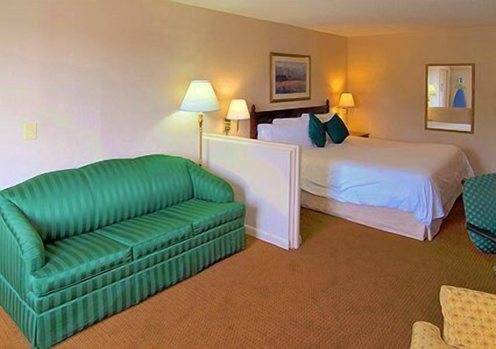 A hotel room with a green sofa, a bed with white bedding, two bedside lamps, and a painting on the wall.