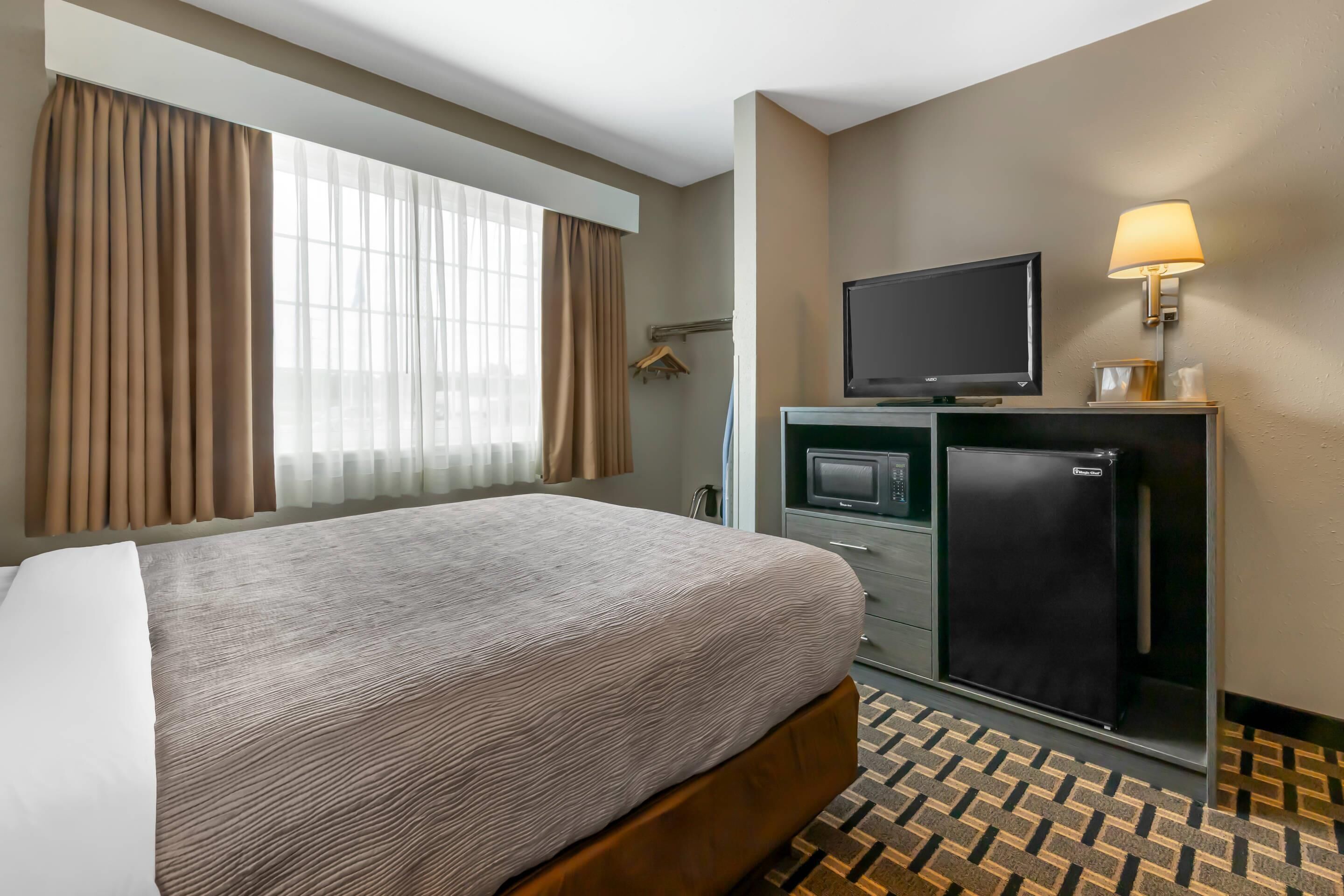 A hotel room with a bed, a television, a refrigerator, and a desk with a lamp.