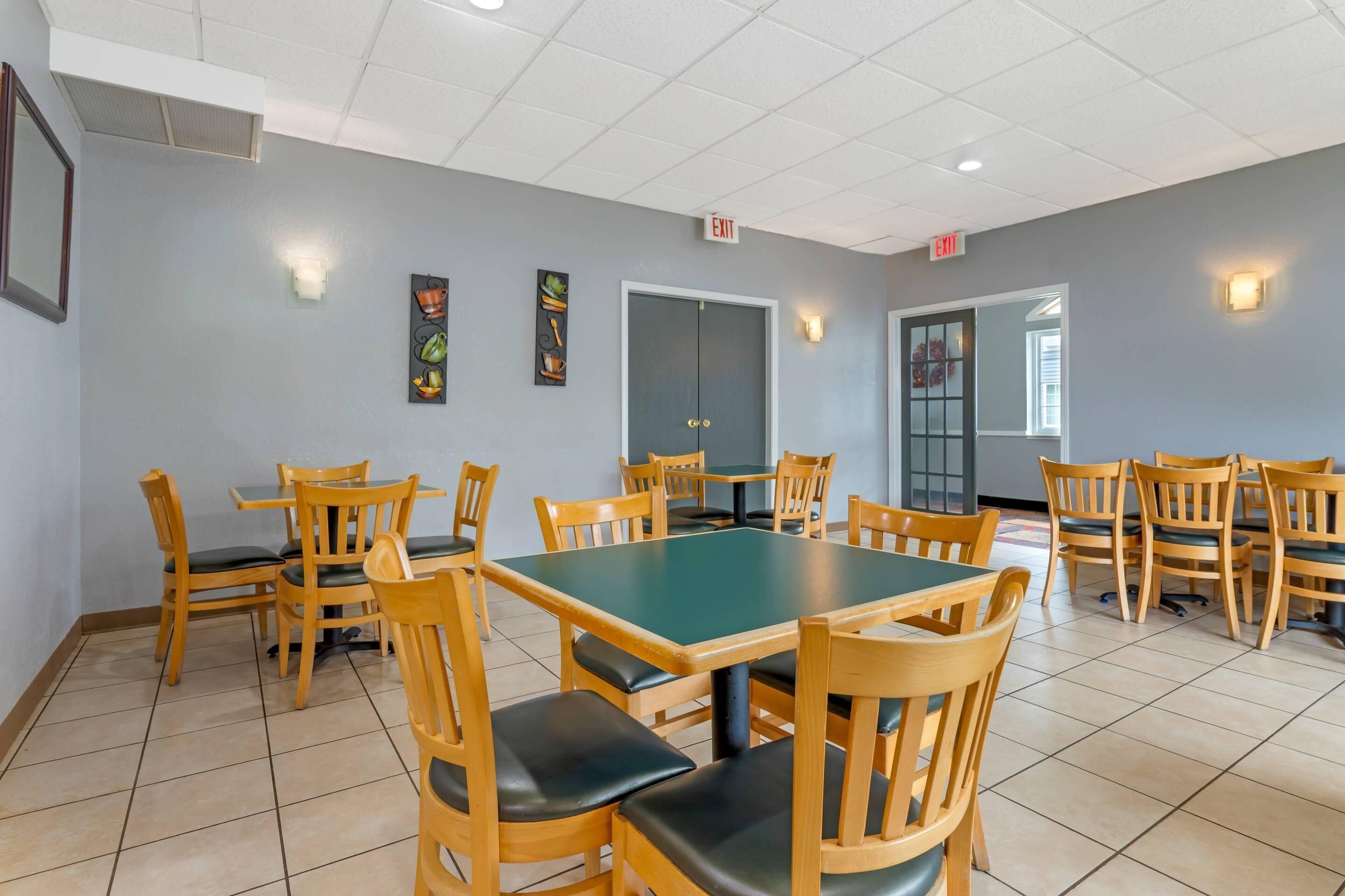 A dining area with multiple wooden tables and chairs, light grey walls, and exit signs.