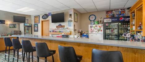 Una zona de bar con mostrador, taburetes, un refrigerador lleno de bebidas y un reloj en la pared.