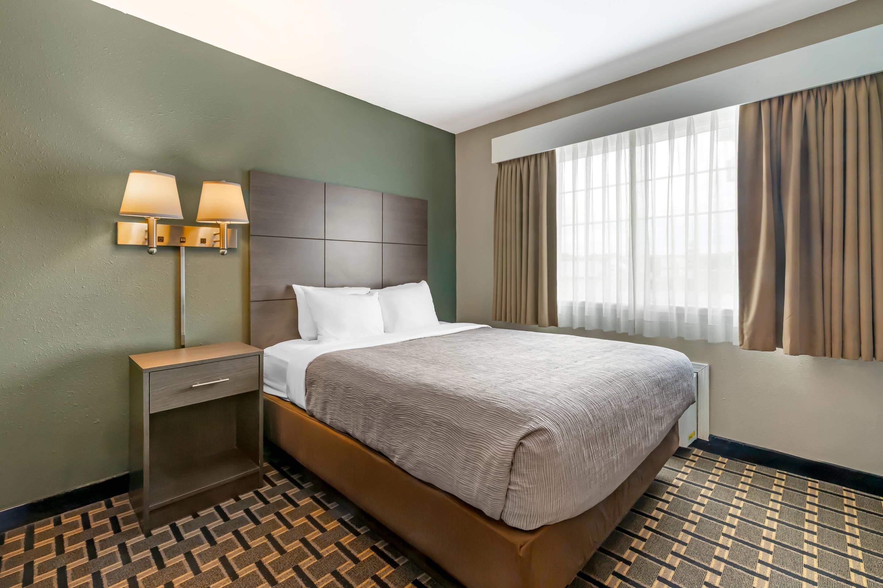 Una habitación de hotel con una cama grande, una mesita de noche con lámparas, un aparador y una ventana con cortinas.