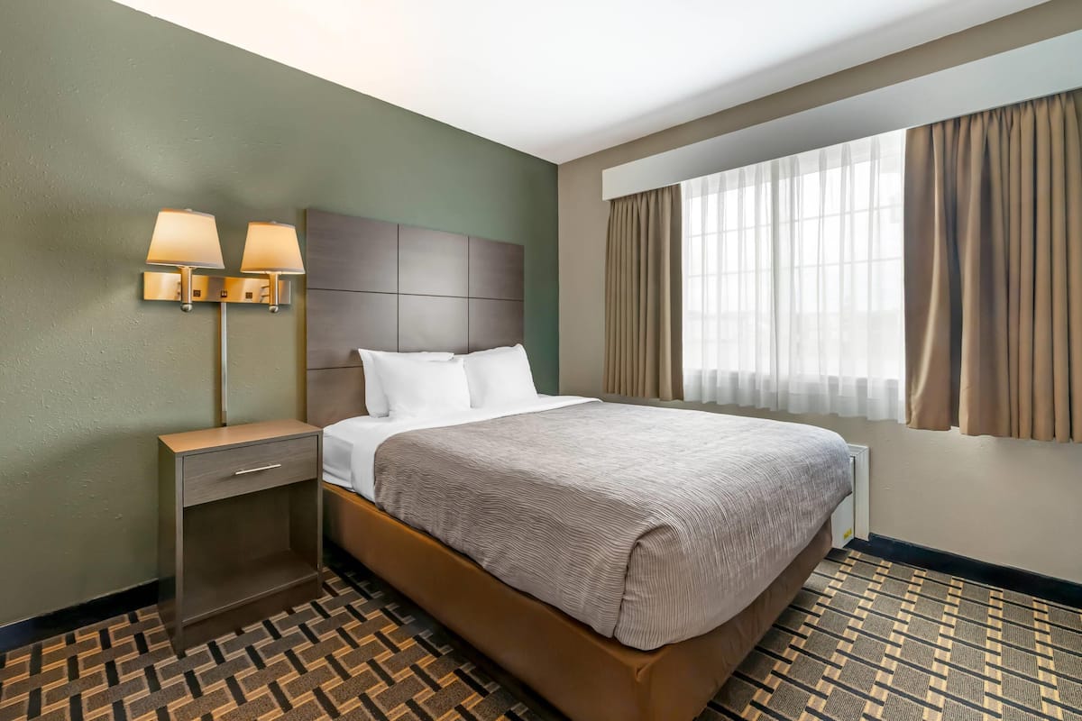 A hotel room with a large bed, a nightstand with lamps, a dresser, and a window with curtains.