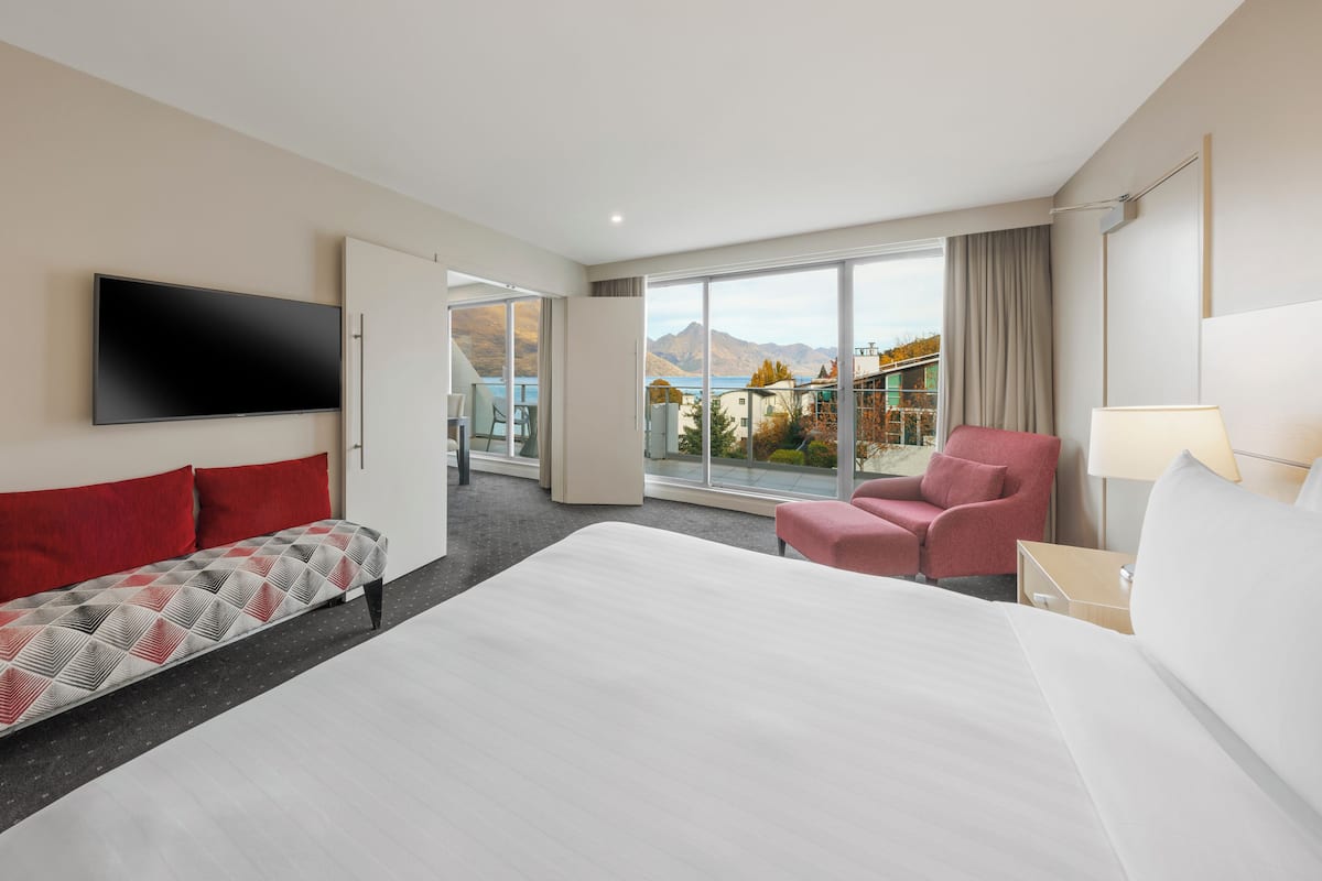 Hotelkamer met een groot bed, een bank, een stoel, een televisie en een balkon met uitzicht op bergen en een meer.