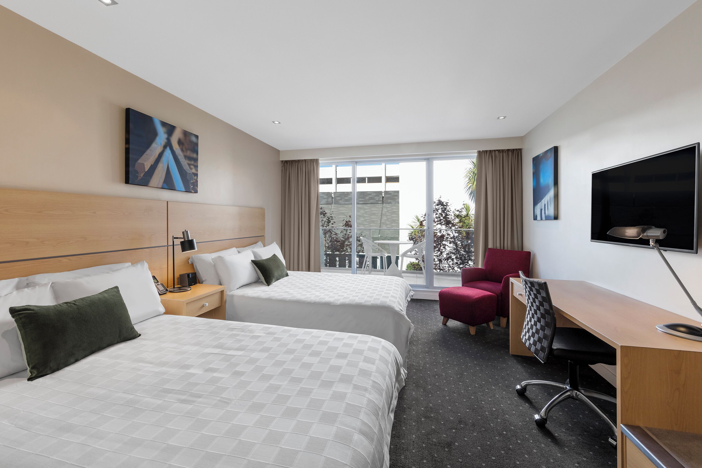 A hotel room with two beds, a desk, a chair, a TV, and a large window.