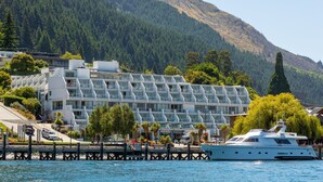 Een modern hotel met een groot zwembad, een steiger met een jacht en een bergachtig landschap op de achtergrond.