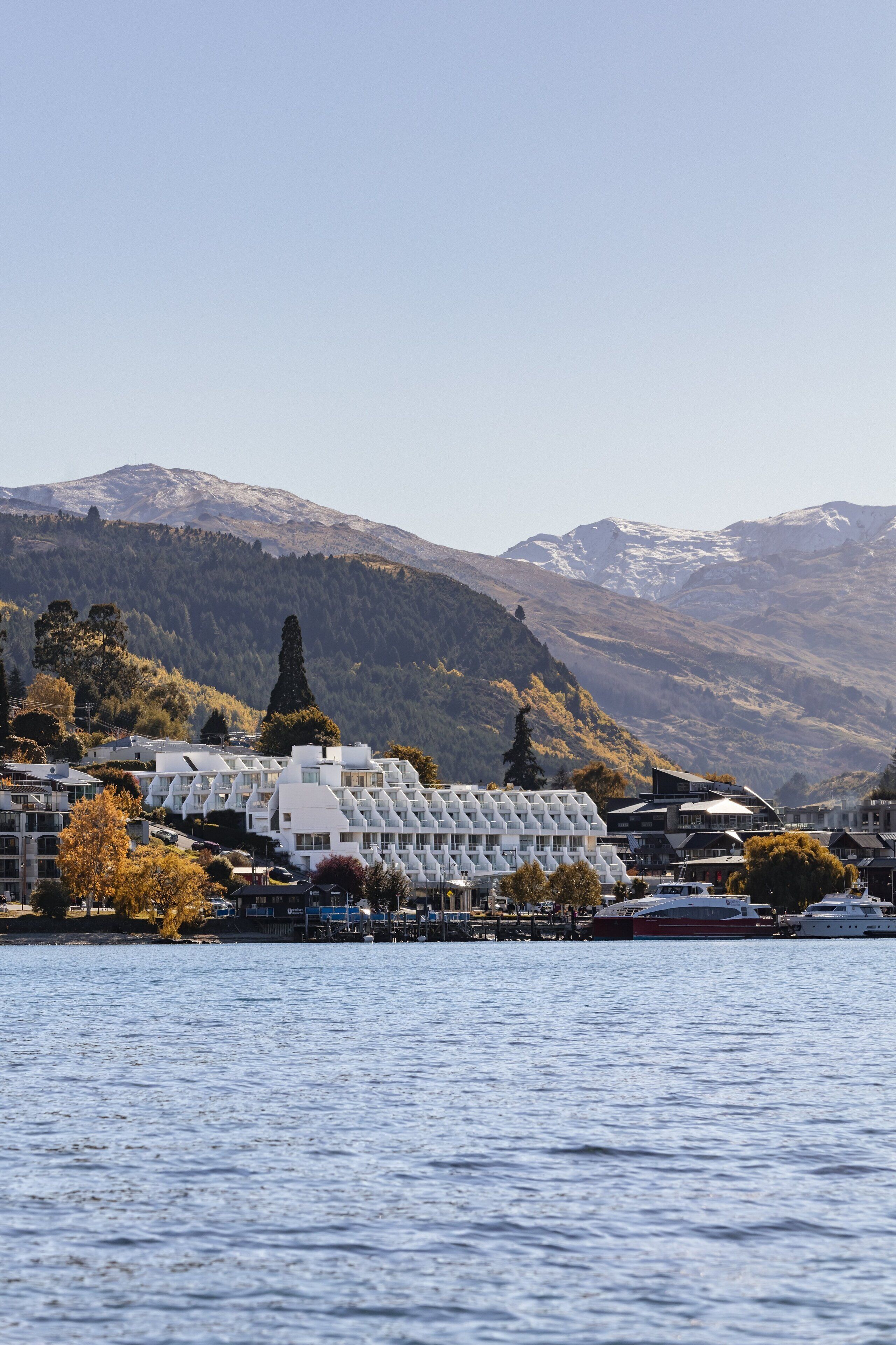 A waterfront hotel with a modern architectural design, located near a lake with mountains in the background.