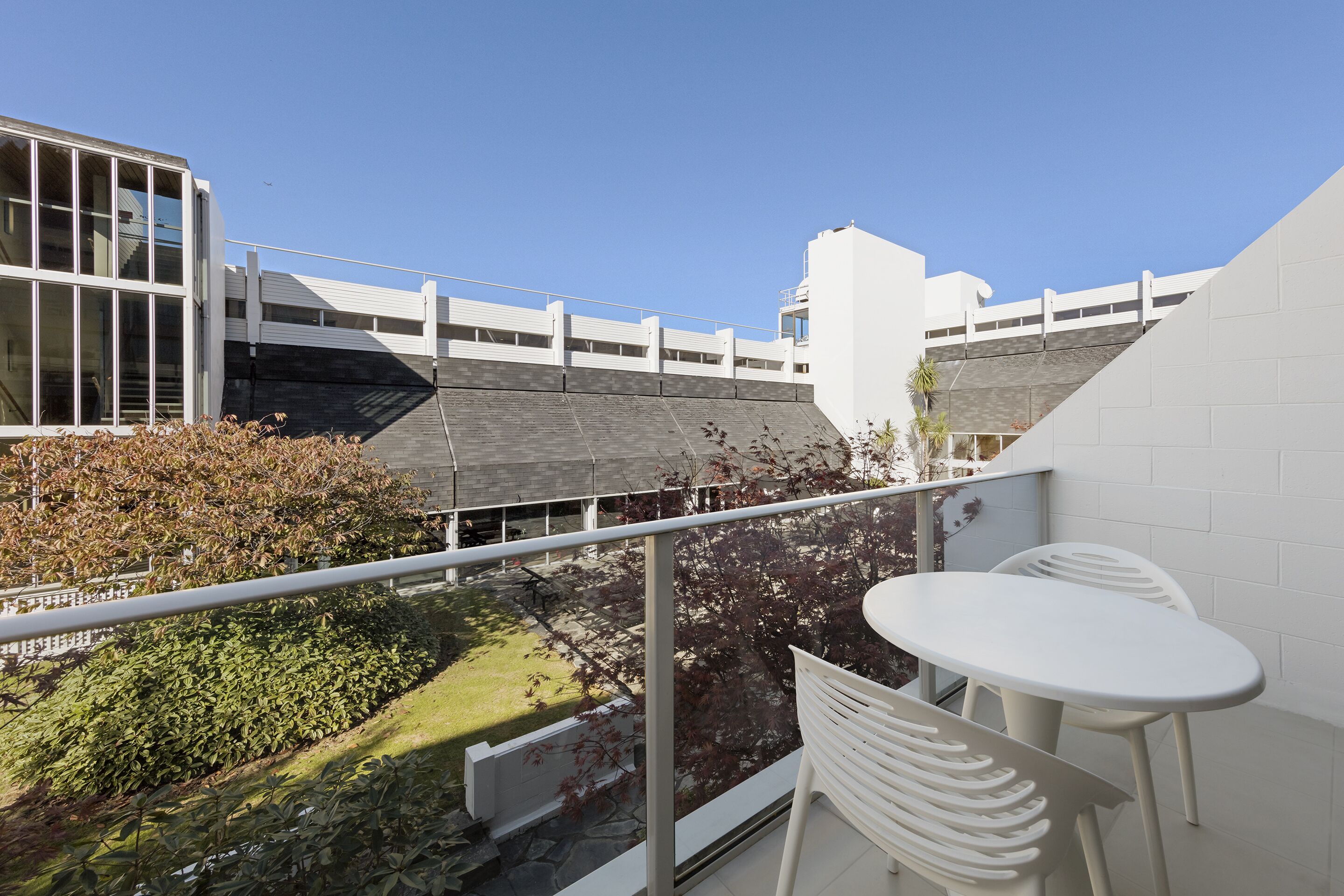 A balcony with a white table and chairs, overlooking a garden and a modern building with large windows.