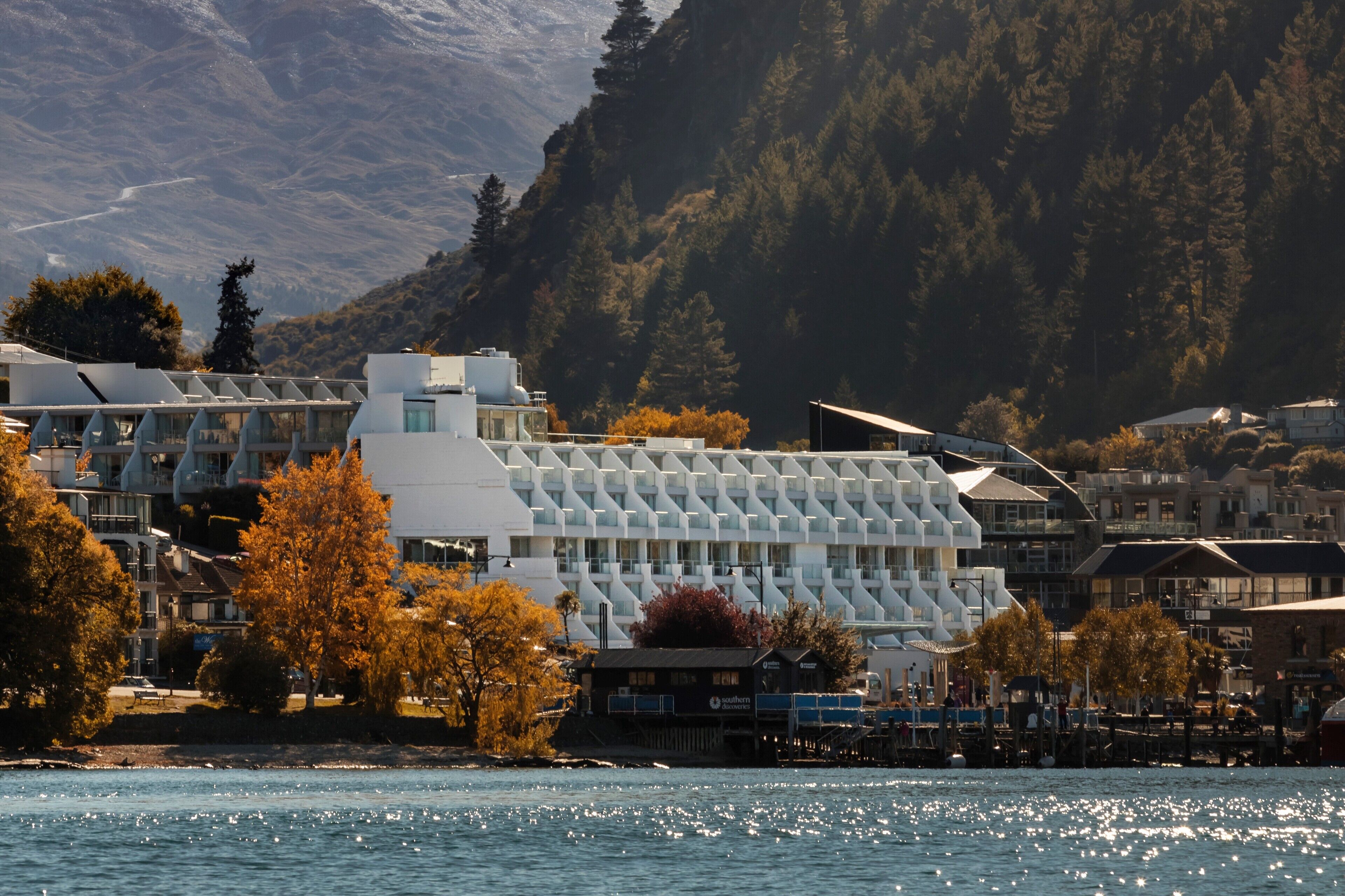 A waterfront hotel with modern architecture, a view of the water, and a mountainous backdrop.