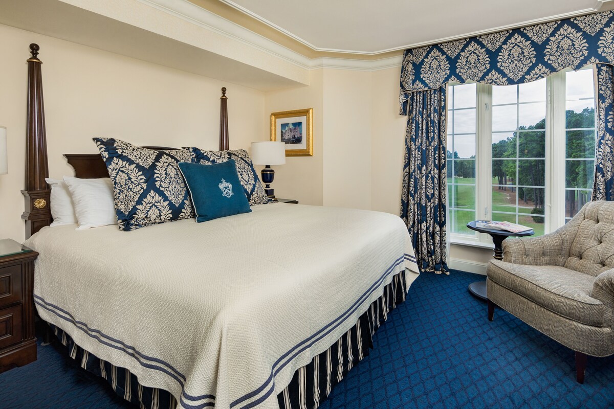 A large bed with a wooden headboard, a small table, and a chair in a room with a blue carpet and patterned curtains.