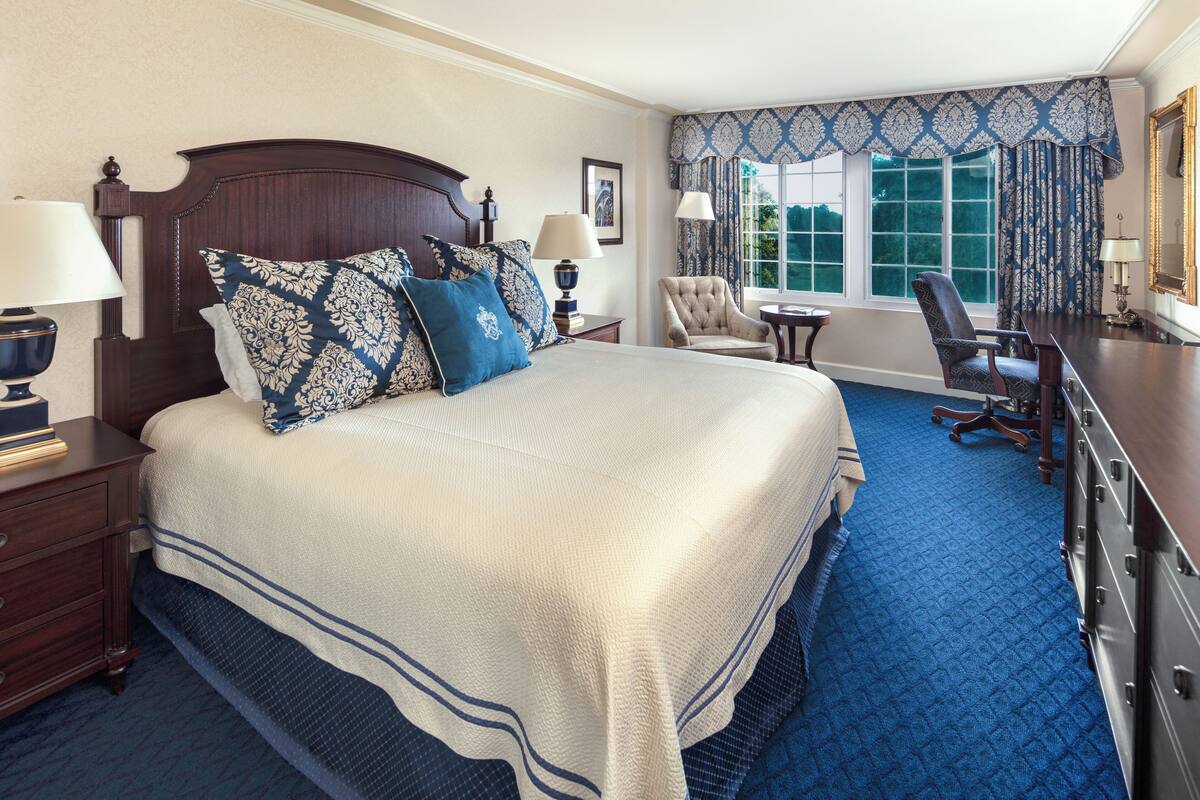 A hotel room with a large bed, two bedside tables with lamps, a chair, a desk, and a window with curtains.