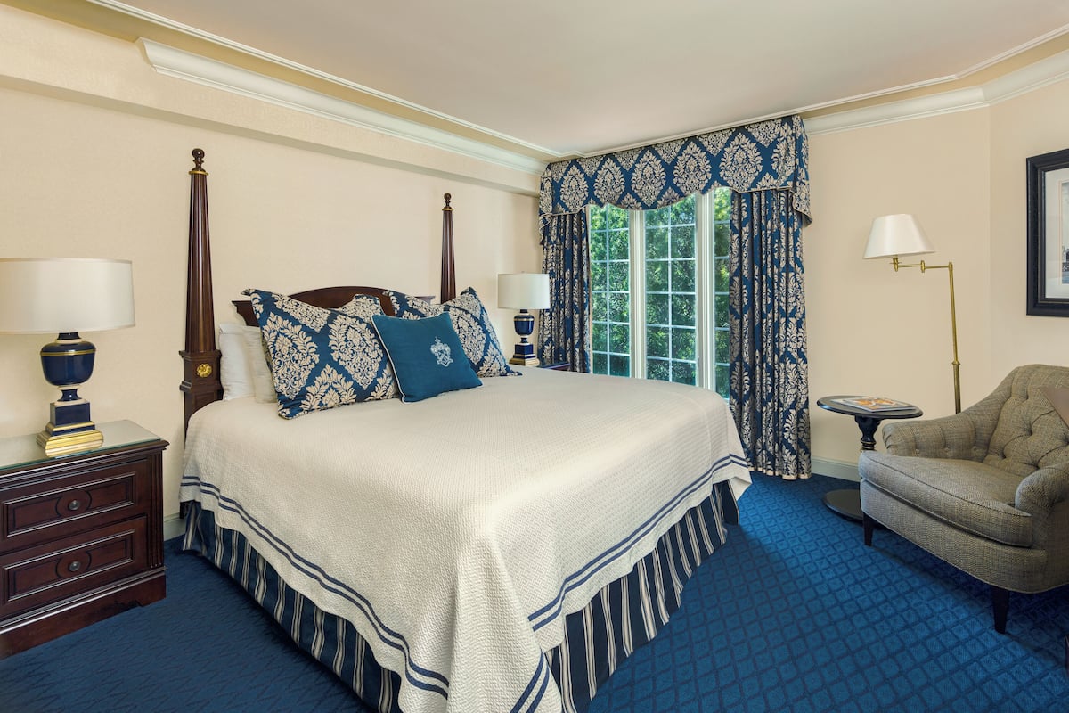 A neatly arranged bedroom with a four-poster bed, a nightstand, a chair, and a window with patterned curtains.