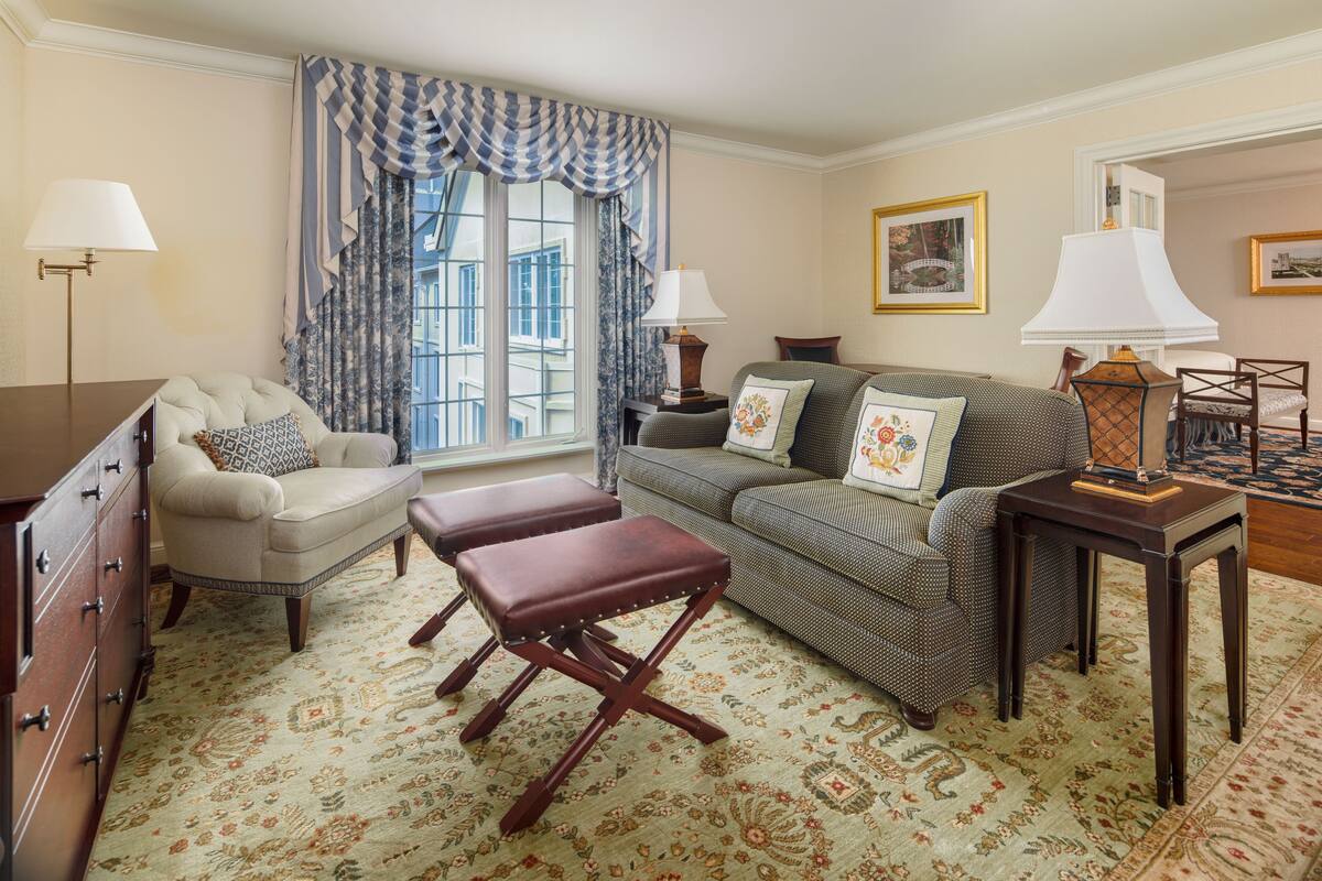A living room with a sofa, armchair, ottoman, and a wooden side table with a lamp.