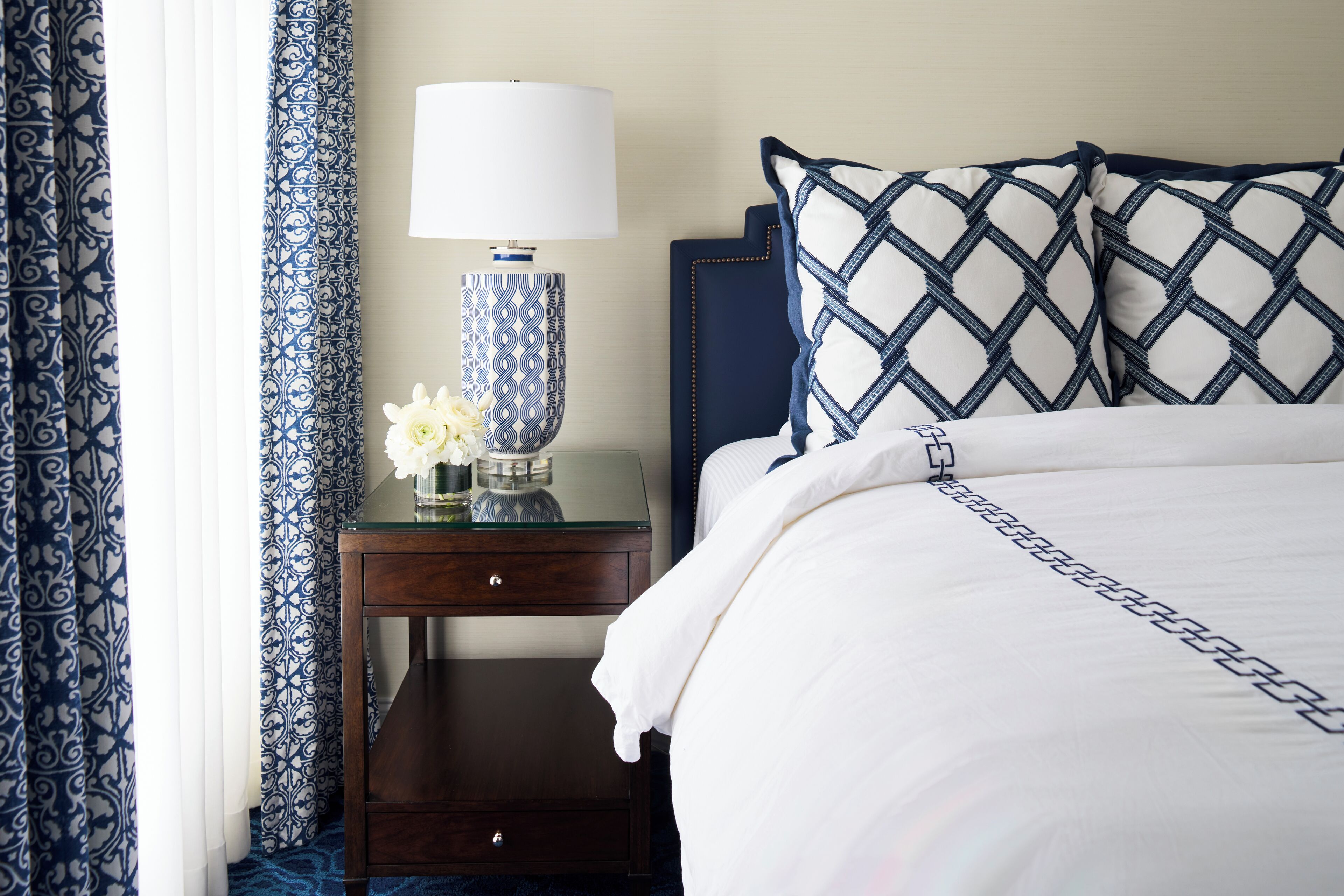 Een net opgemaakt bed met een witte deken en kussens met een blauw-wit patroon, een nachtkastje met een lamp en een bloemenvaas, en een donker houten nachtkastje.