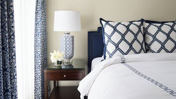 Een net opgemaakt bed met een witte deken en kussens met een blauw-wit patroon, een nachtkastje met een lamp en een bloemenvaas, en een donker houten nachtkastje.