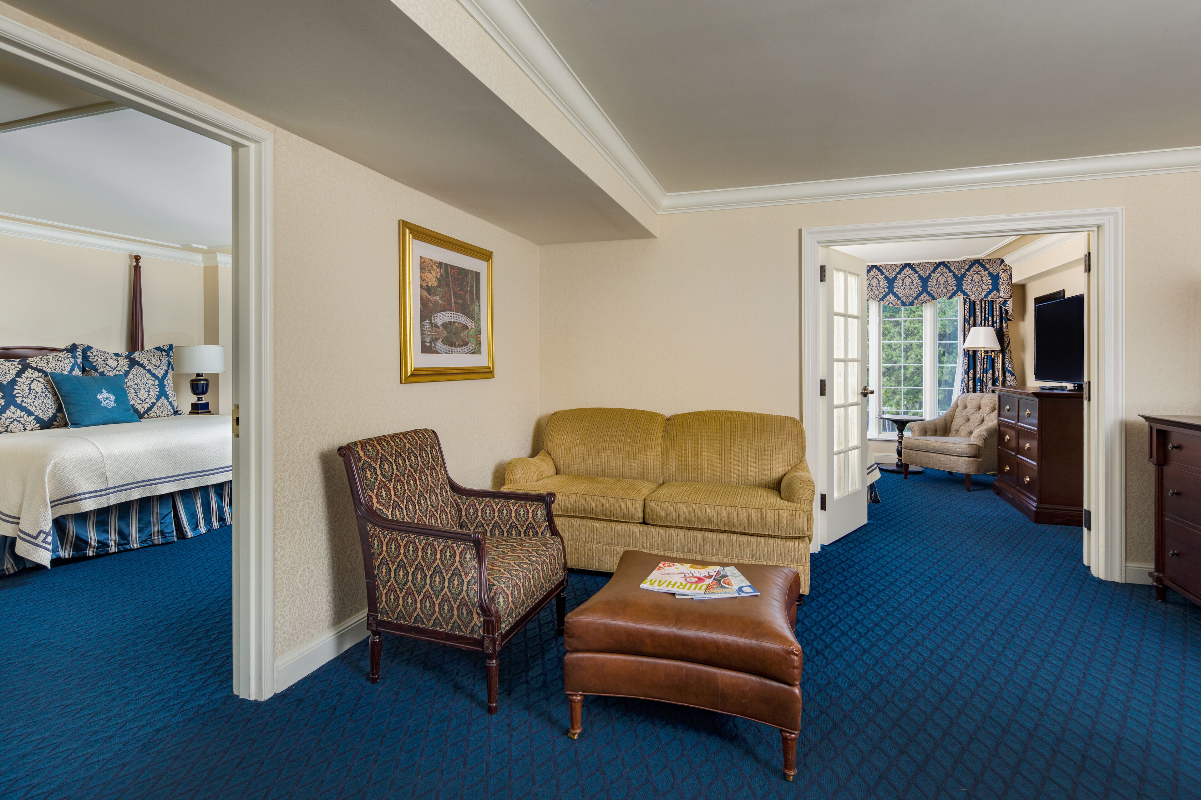 A hotel room with a bed, two sofas, a patterned armchair, a brown ottoman, a dresser, and a television.
