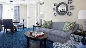 Ein Hotelzimmer mit grauem Sofa, gemusterten Sesseln, einem Esstisch mit Stühlen, einem Couchtisch und einem Spiegel an der Wand.
