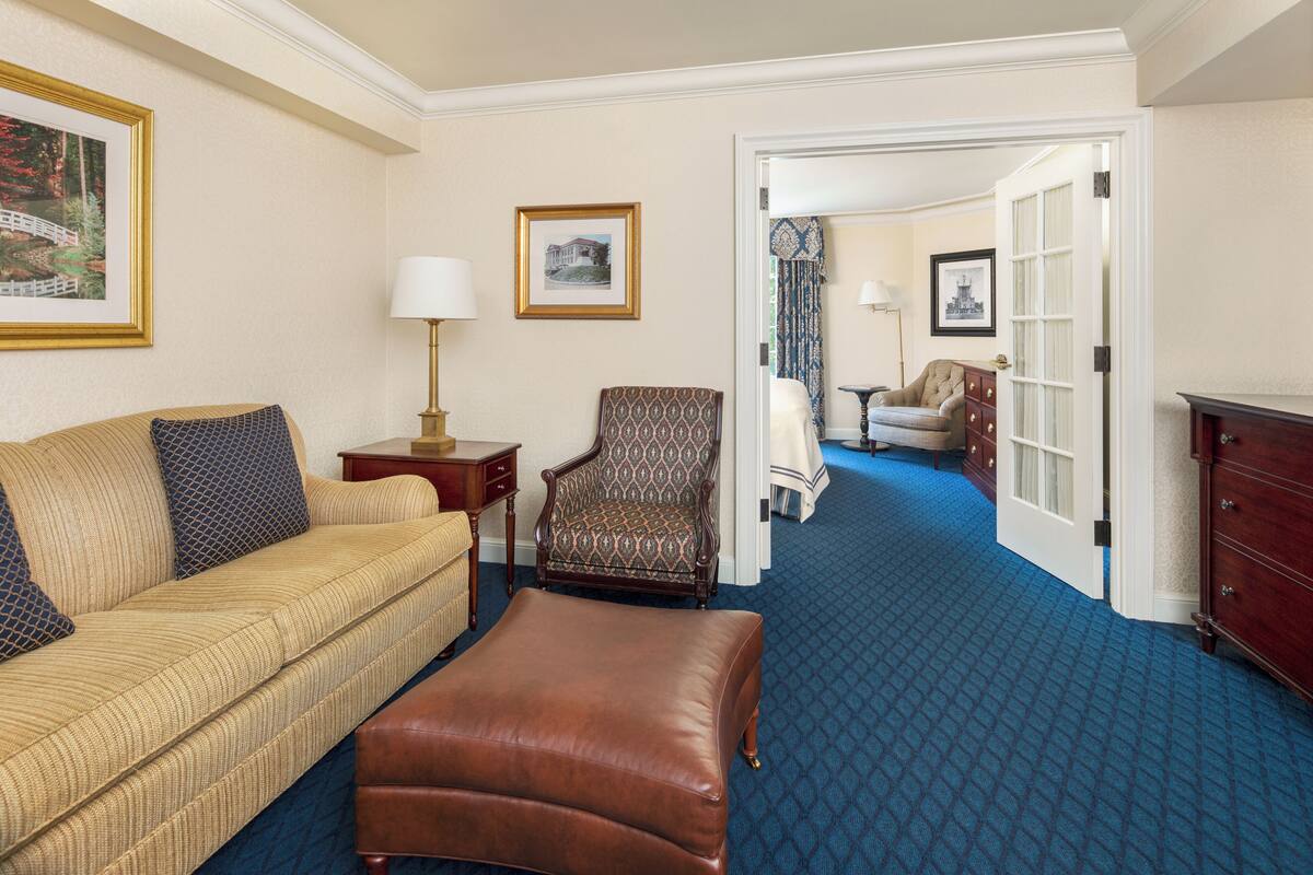 A hotel room with a beige sofa, a patterned armchair, a brown leather ottoman, and a blue carpet.