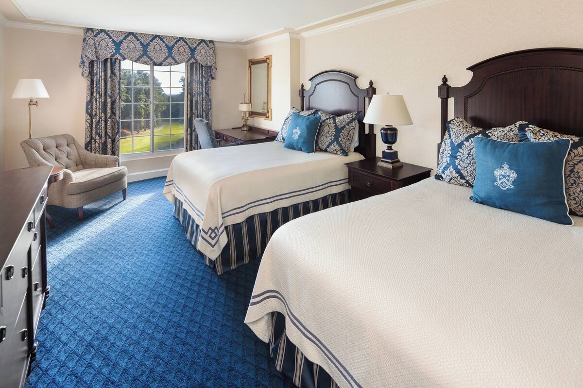 A hotel room with two beds, a chair, a desk, and a window with curtains.