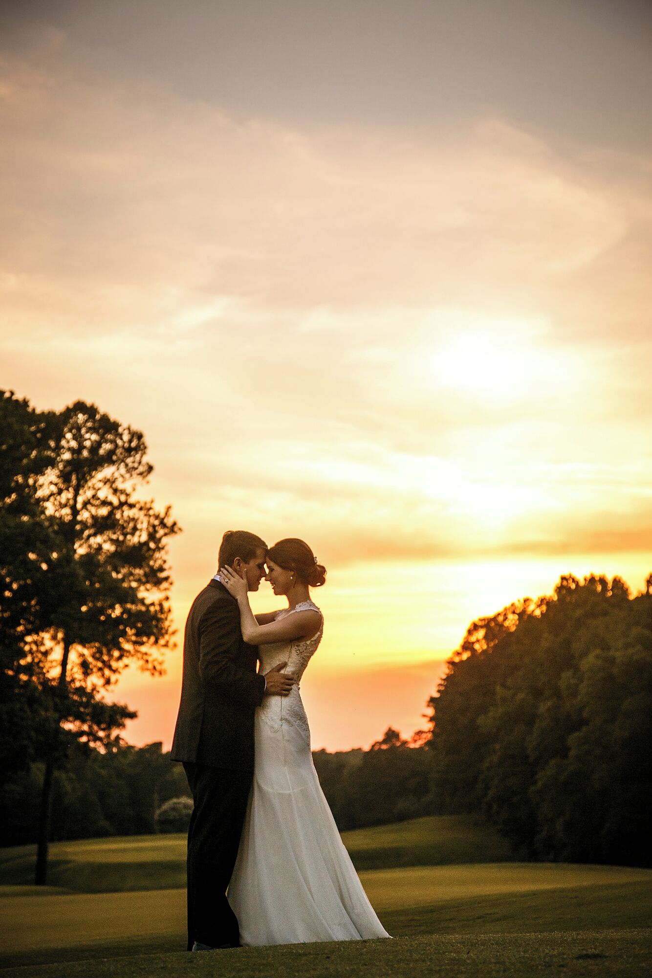 Un couple s’embrasse, la femme portant une robe de mariée et l’homme un costume, et ils se tiennent dans un champ herbeux, avec des arbres et un coucher de soleil en arrière-plan.