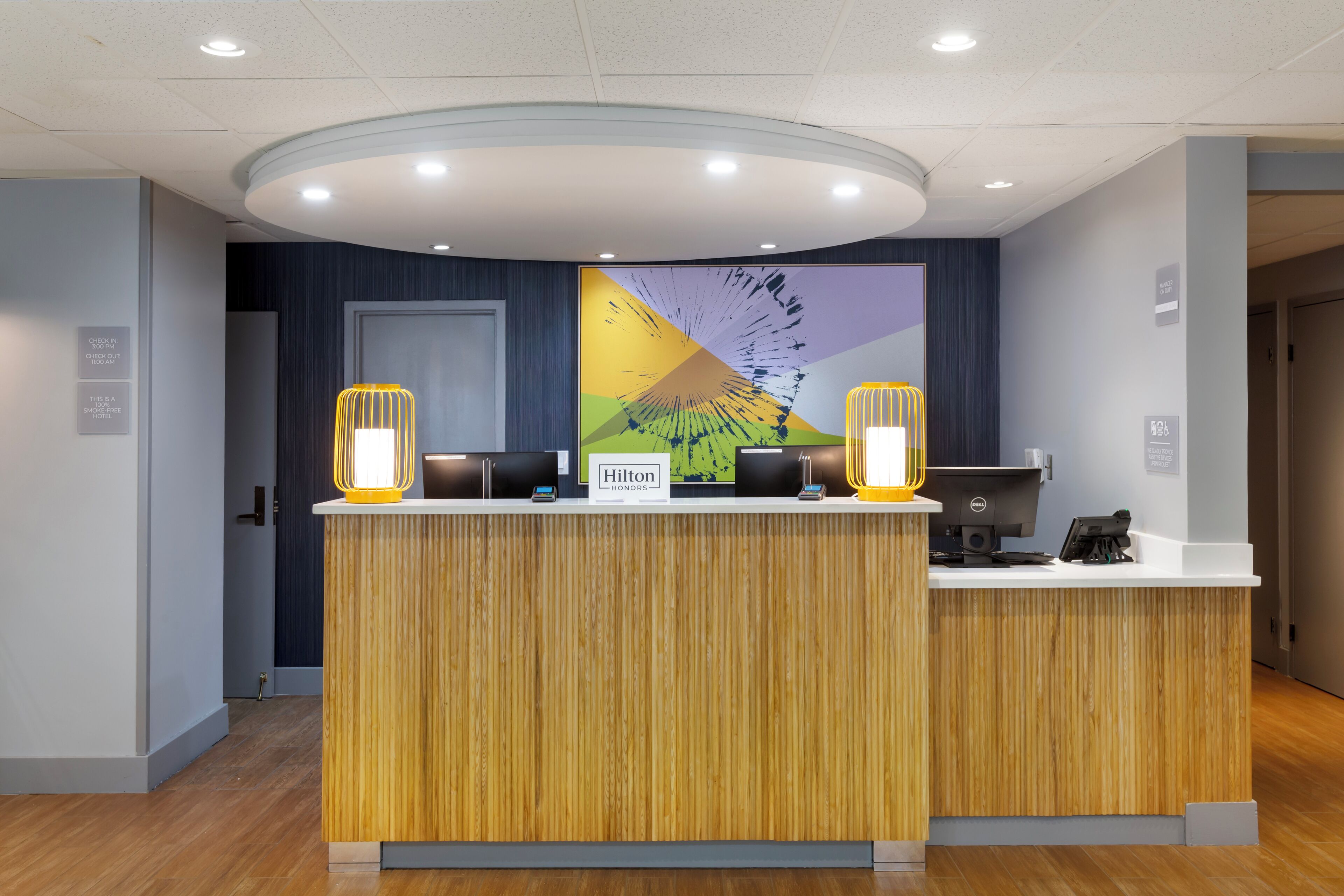 A hotel reception desk with a wooden finish, two illuminated wall sconces, a digital display, and a large abstract painting above.