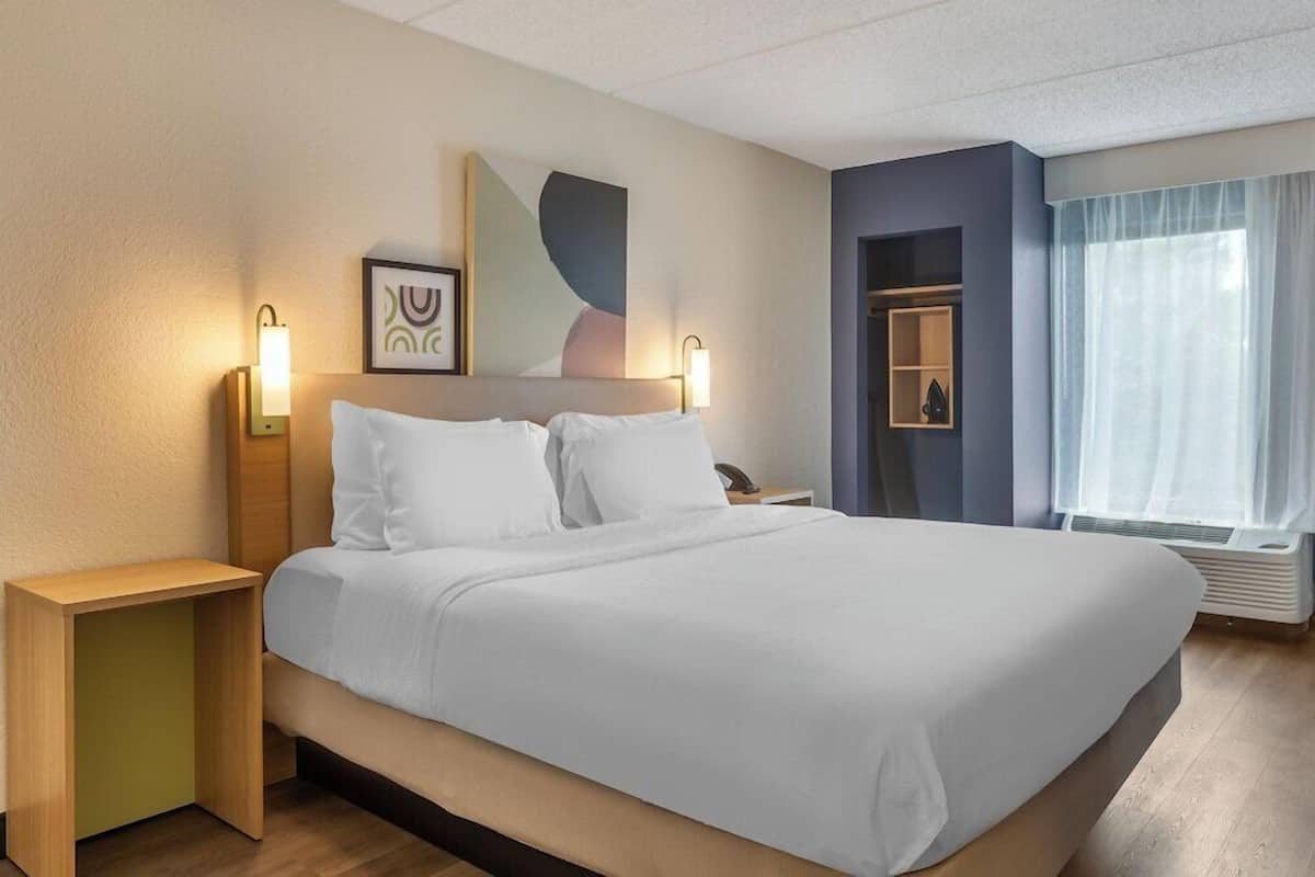 A hotel room with a bed, bedside tables, a wall-mounted artwork, a nightstand, and a window with curtains.