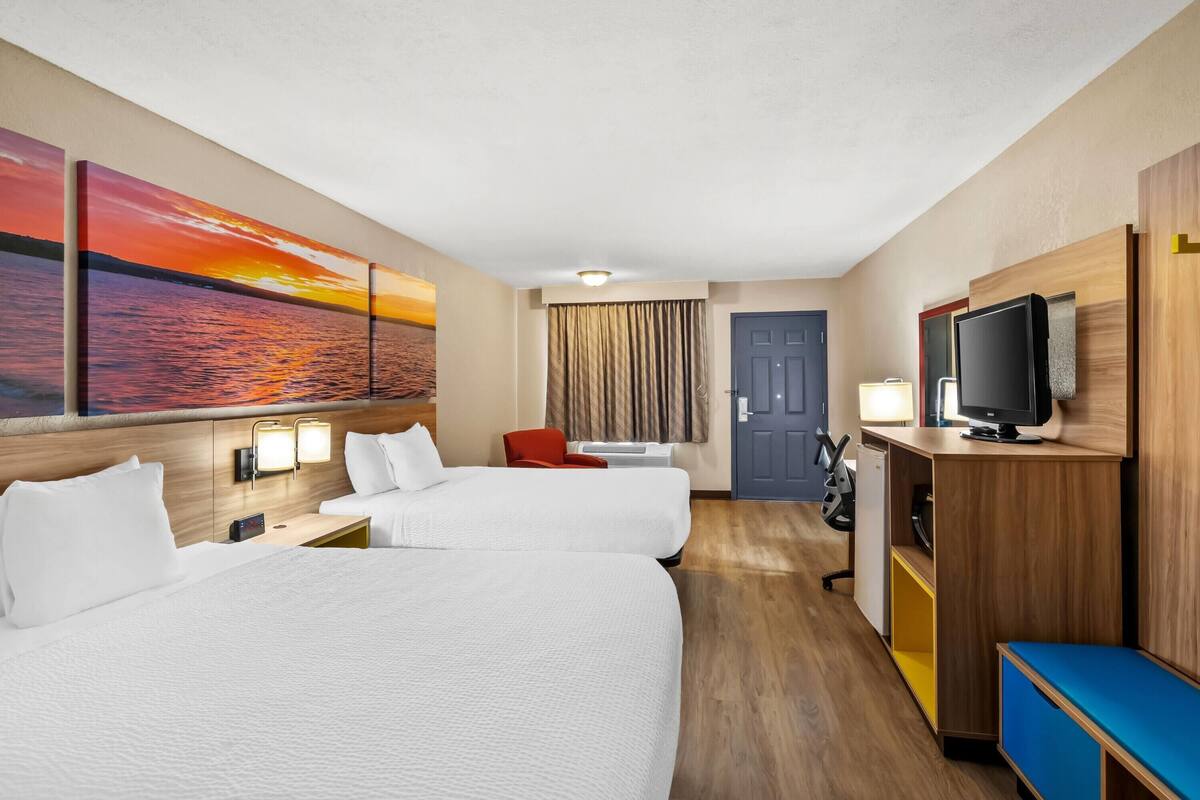 A hotel room with two beds, a TV, a desk, a chair, and a painting of a sunset.
