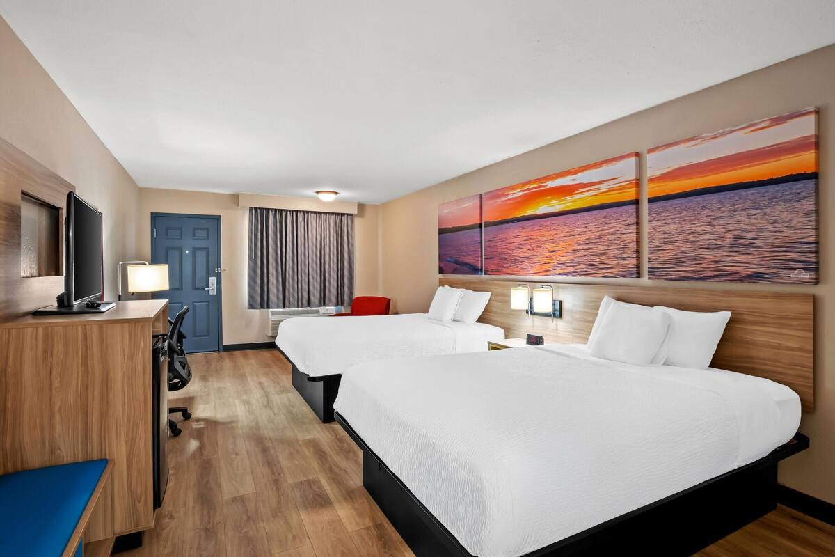 A hotel room with two beds, a desk, a TV, and a painting of a sunset over water.