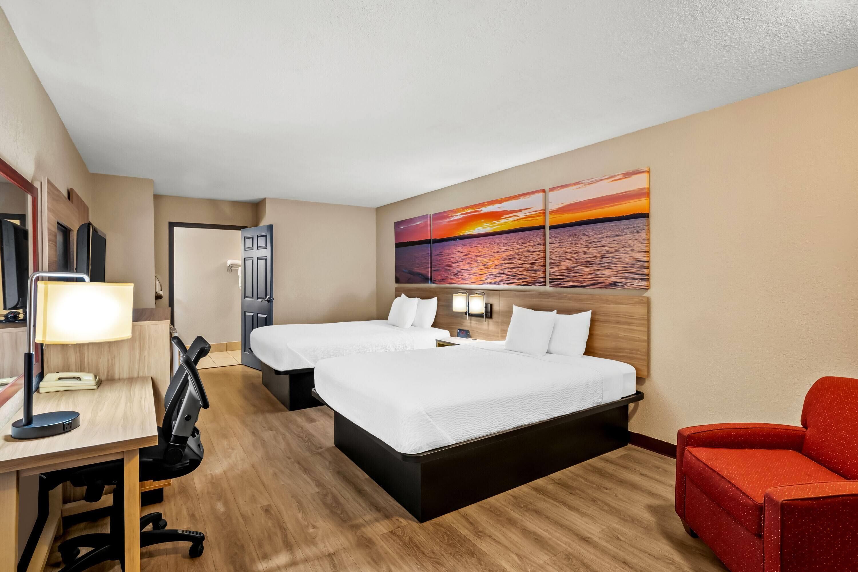 Une chambre d’hôtel avec deux lits, un bureau avec une lampe, une chaise rouge et un tableau représentant un coucher de soleil accroché au mur.