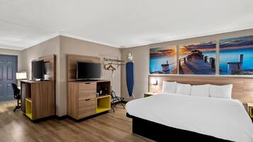 A hotel room with a bed, a desk with a TV, a chair, and a painting of a pier.
