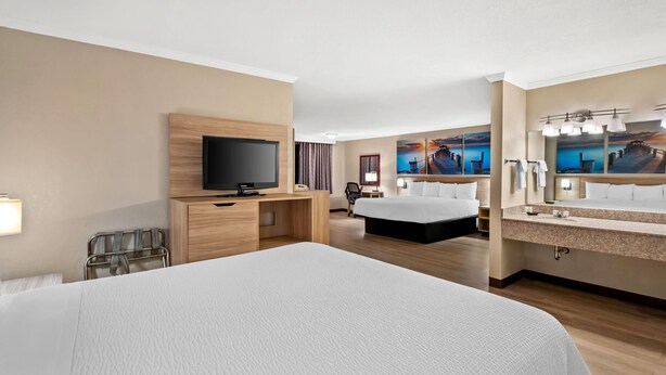 Ein Hotelzimmer mit einem großen Bett, einem Fernseher, einem Schreibtisch, einem Sessel sowie einem Badezimmer mit Waschbecken und Spiegel. An den Wänden befinden sich Kunstwerke.