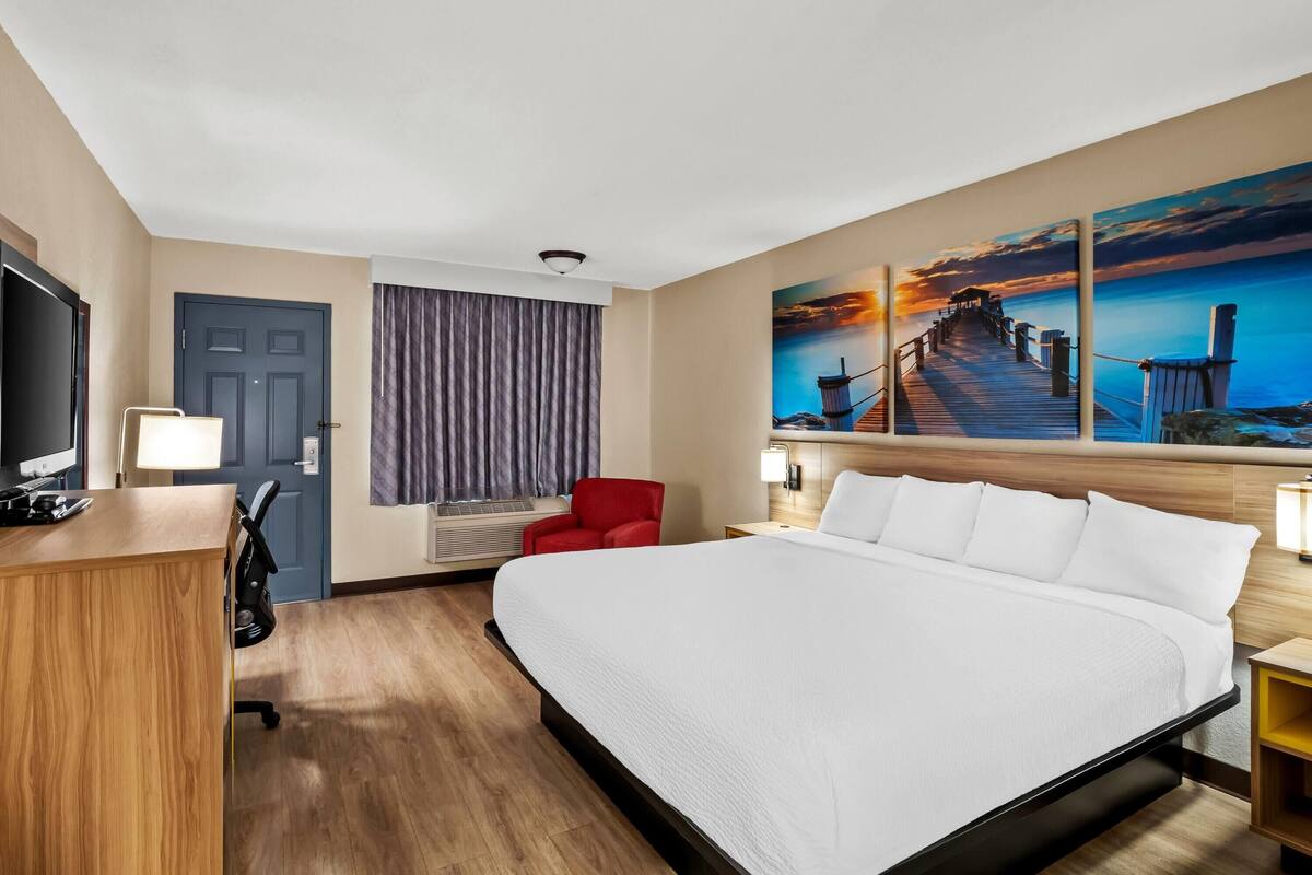 A hotel room with a large bed, a desk, a TV, a red chair, a painting of a sunset, and a view of the sea.