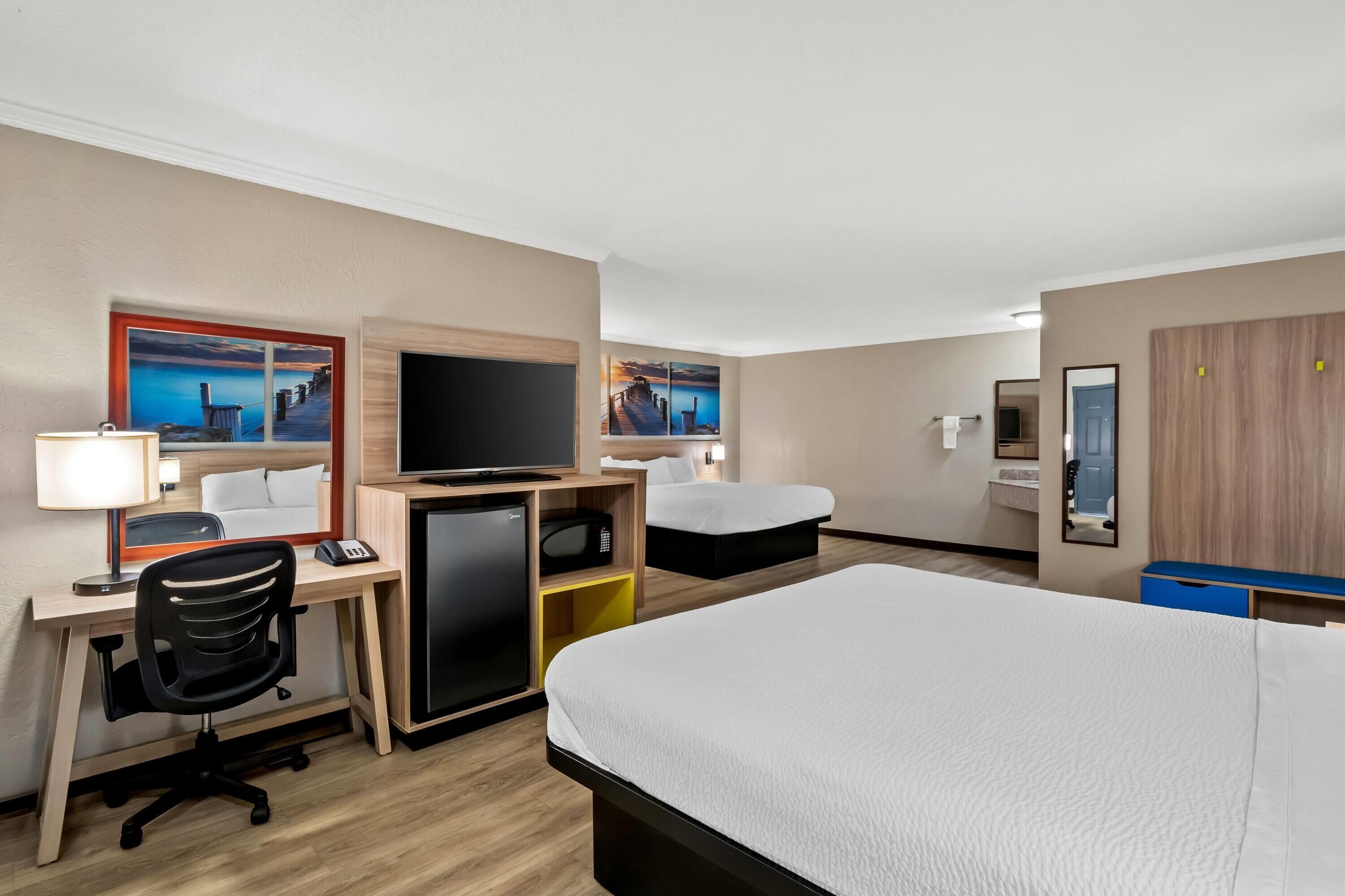 A hotel room with a bed, desk, chair, television, and two paintings on the wall.