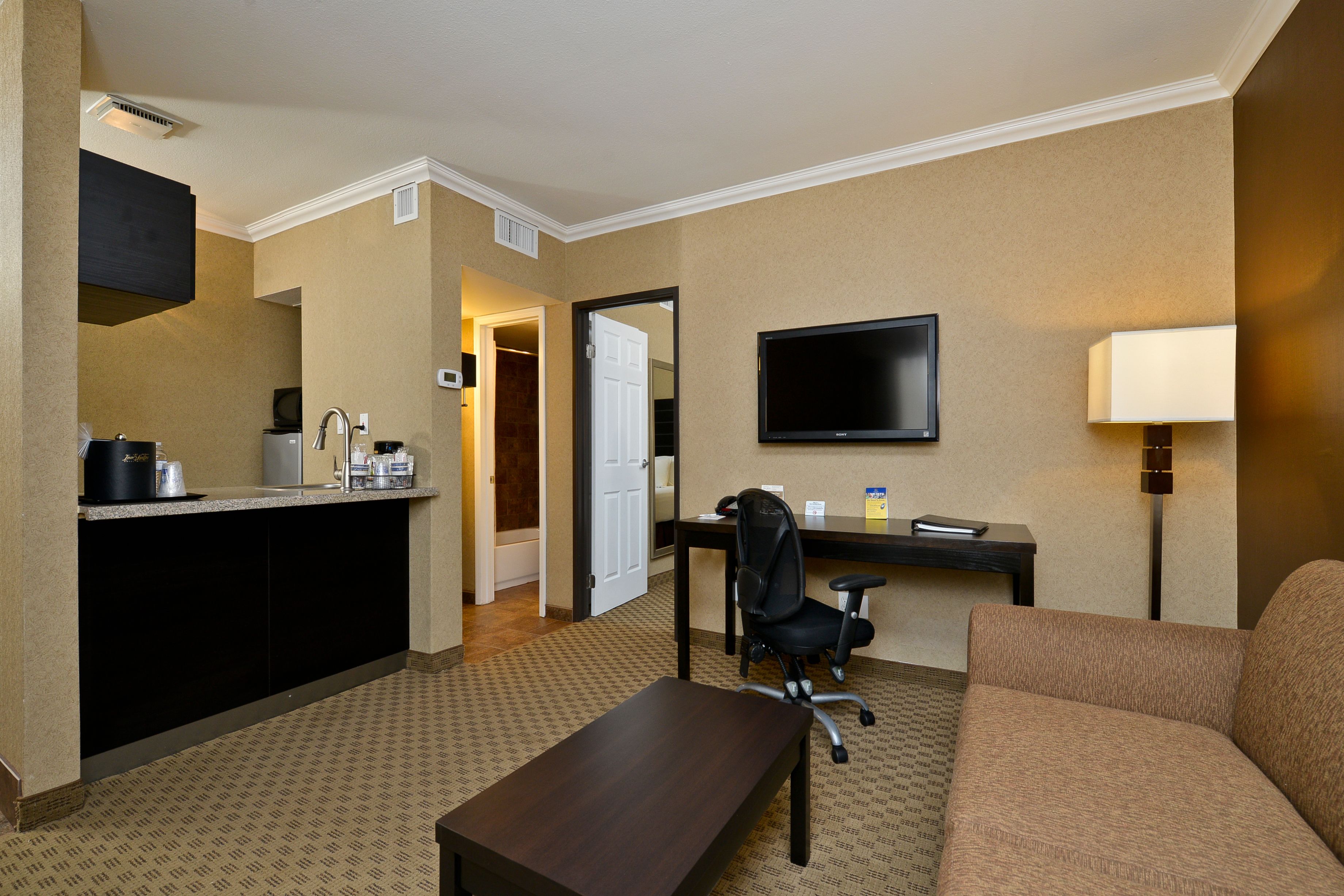 A hotel room with a kitchenette, a flat-screen TV, a desk with a chair, a sofa, and a lamp.