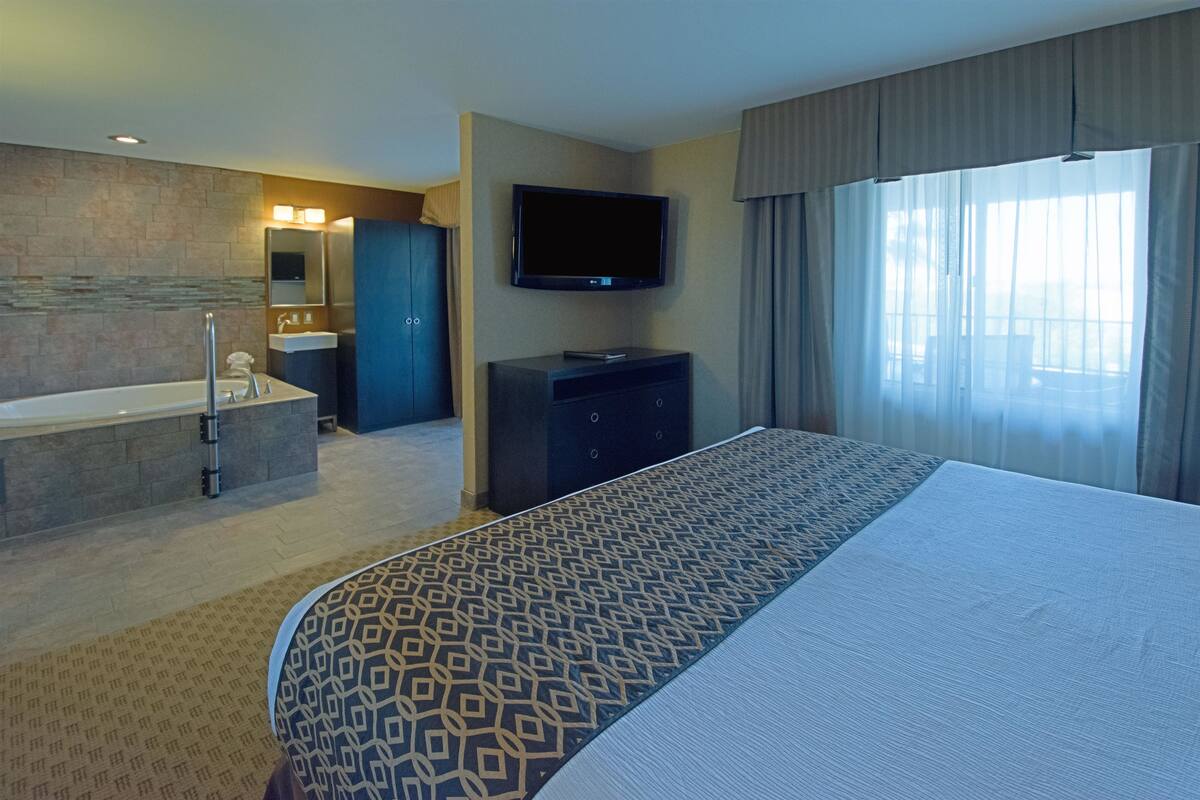 A hotel room with a large bed, a flat-screen TV, a bathtub, a vanity with a mirror, and a large window with curtains.