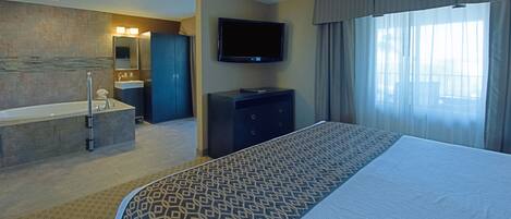 A hotel room with a large bed, a flat-screen TV, a bathtub, a vanity with a mirror, and a large window with curtains.